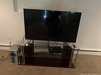 Photo showing a 55 inch Roku flat screen TV on a glass and wood TV stand with a gray subwoofer to the left. A media device with green lights is on the middle shelf and a TV remote rests on top.