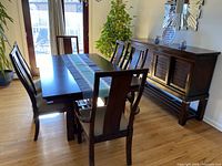 Photo showing dark wood rectangular dining table with 6 matching chairs and buffet sideboard in a well-lit dining area with hardwood floor and wall mirror.