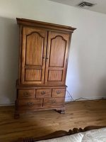 Solid wooden armoire with double doors on top and three drawers below, standing on wood floor near white wall.