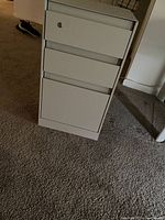 Front and side view of a light gray steel filing cabinet with three drawers on carpeted floor.