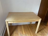 Light-colored wood desk with one drawer, rectangular top and four legs, positioned against white walls on hardwood floor.
