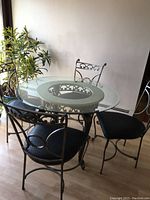 Photo of an oval glass top dining table with decorative black metal base and four matching metal chairs with black cushioned seats.