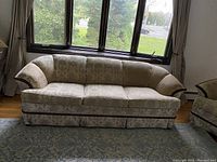 Photo of a three-seat fabric sofa with cover, placed in front of a window with curtains. The sofa has curved armrests, padded backrest, and a skirted base, in fair condition.