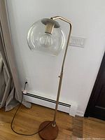 Floor lamp with clear glass globe shade on brass stand and wooden base against a white wall and hardwood floor.