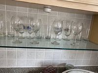 14 clear glasses on a glass shelf, including 6 stemmed wine glasses, 6 beverage glasses, and 2 tumblers.