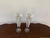 Pair of crystal candle holders with hanging bead strands shown on wooden surface against plain wall.