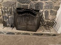 Photo showing a stone fireplace setup with a set of fireplace tools on a stand, a metal fire screen with a curved top, and a metal wood rack for firewood, arranged on a stone hearth.