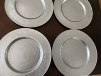 Top-down photo of the 4 round plate chargers laid on a dark wood surface. Each charger has a silver metallic finish with a concentric textured pattern.