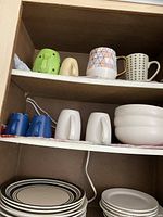 Photo of cabinet shelf with four white dinner plates, four white bowls, two blue mugs, two white mugs on the middle shelf, and an upper shelf with four patterned mugs in various designs including green, beige, pink geometric, and white with black dots.