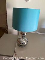 Single table lamp placed on a white surface against a plain wall. Lamp has a chrome glossy spherical base with a blue cylindrical shade.