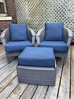 Front view of two outdoor wicker chairs with blue cushions and a matching ottoman, showing the set arranged on a wooden deck with lattice background.