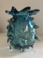 Front view of small round blue glass vase with multiple thick pointed spikes covering the surface, placed on a white surface with plain background.