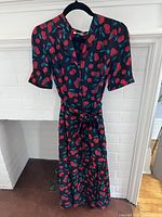 Full length front view of midi dress with cherry print hanging on a black hanger against a white brick wall