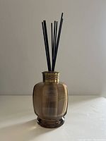 Front view of smoky glass diffuser bottle with black diffuser sticks inserted, sitting on white surface with plain background.