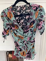 Front view of blouse showing large floral pattern in pink, orange, and black on turquoise base with elbow-length ruched sleeves.
