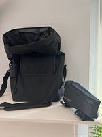 Two black bike bags placed side by side on a shelf, Arkel Signature V Large Bike Bag on left with roll-top open and Blackburn Outpost Top Tube Bag 2.0 on right.