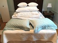 Photo showing a stack of six white pillows, white quilts, white sheets, white bed skirt, and two light blue quilted cushions on a bed.