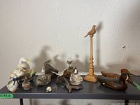 Direct front view of all bird figurines displayed on a shelf including wooden pedestal bird, painted birds on rocks, duck decoys, and small carved birds.