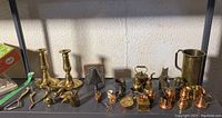 Shelf with brass candlesticks, copper and brass pitchers, teapots, a hand bell, miniature coffee grinder, and small figurines.