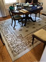 Overall view of the carpet under a wooden dining table and chairs in a room with hardwood flooring, showing its floral and ornamental border pattern in beige and cream colors.