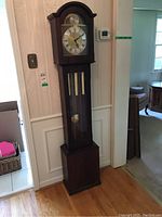 Photo of a tall grandfather clock against a wall showing wooden case, glass front, metal weights and pendulum