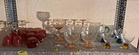 Shelf view showing various cranberry and clear glassware including stemmed glasses and cups arranged in row.