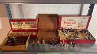 Wide view of multiple boxes with soldier figurines in red and brown Britains boxes on shelf.