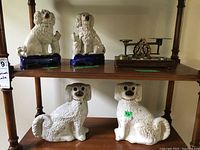 Four antique white ceramic dog figurines and one wooden & brass postal scale displayed on two shelves.