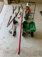 Overview of entire gardening tools lot including the Scotts Turf Builder spreader, electric chainsaw, hedge trimmer, multiple hand tools, watering can, and terracotta pots on concrete floor.