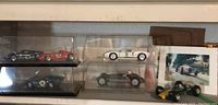 Shelf with four clear plastic display cases containing five race car models, one loose green-and-yellow open-wheel car, and a printed racing car card.