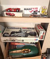 Two-tier shelf showing assembled red Lola T70 on its box above a Tamiya Brabham BT44B box and green hydroplane hull below, with stickers behind.