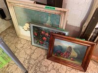 Three paintings grouped together, including one framed basket of strawberries painting and two other still lifes. Frames show some damage.