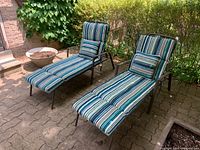 Two metal frame patio loungers with striped cushions and adjustable backs are placed on a paved outdoor patio near greenery and planters.