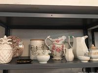 Shelf display of the collection showing multiple antique pitchers, cream and sugar sets, a ceramic canister with butterfly design, Moorcroft trinket dish, pink floral plate, milk glass vase, and a pineapple jar.