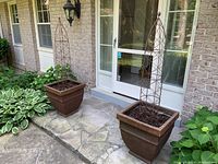 View of two square resin planters with rustic brown finish placed on outdoor patio with foliage nearby, showing overall look and size.