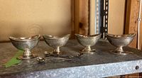 Four silver pedestal bowls arranged in row with three spoons in front on metal shelf