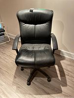 Front view of black leather office desk chair showing cushioned seat and back, fixed armrests, and five-wheeled pedestal base.