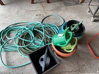 Coiled green and beige garden hoses laid on the ground beside a stack of plastic watering cans and planter pots. Includes black square planter, terracotta colored round planter, and multiple blue round planters.