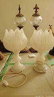 Pair of white flower-shaped table lamps with electrical cords visible, placed on a table along with other lamps.