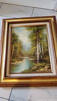 Framed oil painting showing a landscape of stream and trees, signed in lower left, in a gold-tone wooden frame.