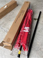 New Smirnoff branded patio market umbrella canopy in red, wrapped in plastic, alongside black metal frame poles and original cardboard packaging boxes on the floor.