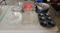 Clear glass Pyrex bowls, a rectangular glass baking dish, a red-labeled baking pan, metal cake pans and muffin tin, and a springform pan arranged on a table