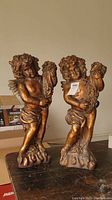 Front view of two bronze-finished cherub statues standing on bases, holding floral wreaths, displayed on a table.