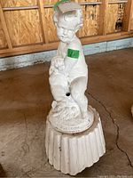 View of statue of a naked child standing on a round fluted base with figurine in hand, showing front and left side details.