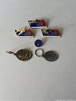 Photo showing three Wings of Victory enamel pins featuring golden airplanes above patriotic backgrounds, one round blue Wings of Victory pin, and two oval National Championship Air Races keychains from 1995 (bronze) and 1998 (silver).