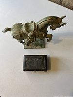 Photo showing a vintage bronze or brass horse statue with a greenish patina, and a small rectangular Greek souvenir box with an embossed temple image beside it.