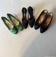 Three pairs of women's designer heels displayed side by side on a plain surface: one green pair with bows, one black pair with decorative buckles, and one plain black pair.