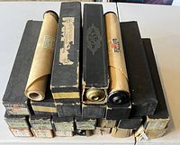 Photo showing 24 player piano rolls stacked in original black boxes and cylindrical cardboard tubes with some boxes worn and aged.