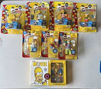 Photo showing all figures from World of Springfield series (Nelson, Moe, Otto, Milhouse), talking clip-on figures (Hungry Homer, Slurpin Bart, Krusty the Clown), and The Simpsons movie DVD with five figures included, with visible wear on boxes.