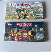 Photo showing two unopened Monopoly board games themed after The Simpsons TV show. One box depicts the Springfield edition with colorful artwork of characters and locations from the series. The other shows the Treehouse of Horror Collector's edition with Halloween themed graphics and characters in costumes.